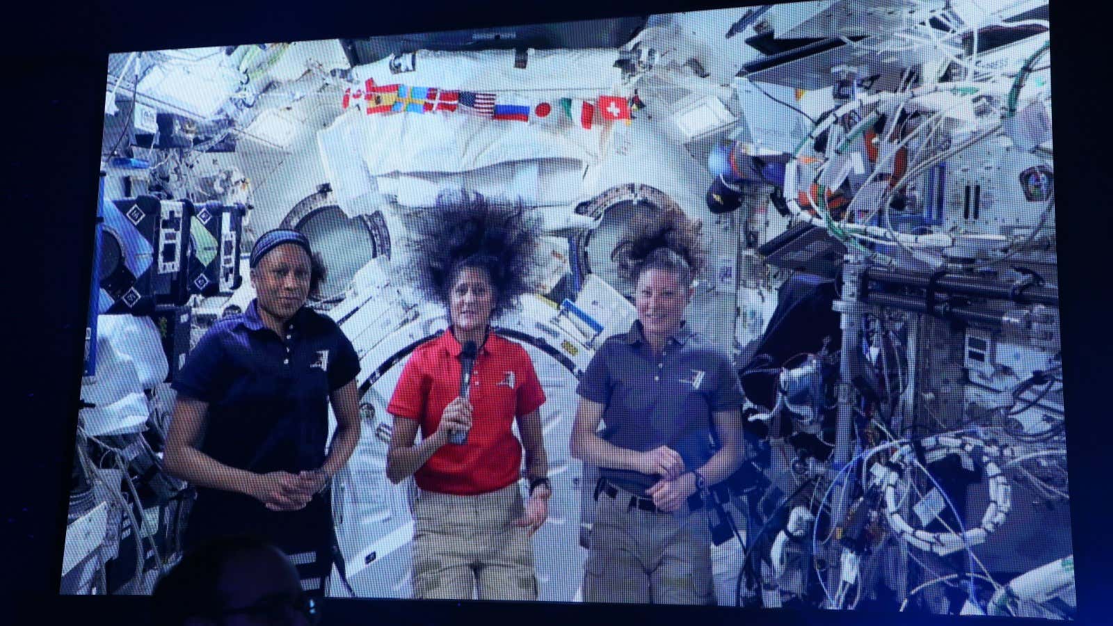 A photo of a monitor showing three astronauts on the International Space Station. 